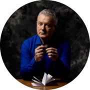 Ein Mann mit kurzen grauen Haaren und einem hellblauen Hemd sitzt an einem Tisch, hält ein aufgeschlagenes Notizbuch und einen Stift in der Hand und blickt nachdenklich in die Kamera vor einem dunklen, strukturierten Hintergrund.