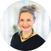 Eine lächelnde Frau mit hochgesteckten hellen Haaren, die eine klobige grüne Perlenkette und ein schwarzes Oberteil trägt, steht in einem hell erleuchteten, verschwommenen Innenraum.