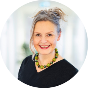 Eine lächelnde Frau mit hochgesteckten hellen Haaren, die eine klobige grüne Perlenkette und ein schwarzes Oberteil trägt, steht in einem hell erleuchteten, verschwommenen Innenraum.