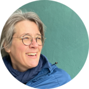 Eine Person mit kurzen grauen Haaren und Brille lächelt und schaut zur Seite. Sie trägt eine blaue Jacke und einen dunklen Schal und steht vor einer schlichten grünen Wand.