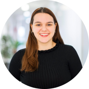 Eine junge Frau mit langen braunen Haaren, die ein schwarzes Oberteil trägt, lächelt in die Kamera. Der Hintergrund ist mit weißen Wänden und einem Hauch von Grün sanft verschwommen. Das Bild ist mit einem kreisförmigen Ausschnitt gerahmt.