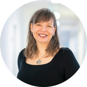 Eine Frau mit schulterlangen braunen Haaren und Pony, die ein schwarzes Oberteil und eine silberne Halskette trägt, lächelt in einem hell erleuchteten Flur in die Kamera.