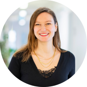 Eine Frau mit langen braunen Haaren, die ein schwarzes Oberteil mit Spitzenverzierung und mehrlagigen Goldketten trägt, lächelt in die Kamera. Der Hintergrund ist in hellen Tönen sanft verschwommen.