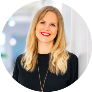 Eine Frau mit langen blonden Haaren, die ein schwarzes Oberteil und eine Halskette trägt, lächelt, während sie in einem Haus vor einem hellen Hintergrund steht.