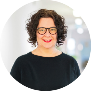 Eine Frau mit lockigem dunklem Haar, schwarzer Brille und schwarzem Oberteil, lächelt in die Kamera. Der Hintergrund ist sanft verschwommen und hell beleuchtet.