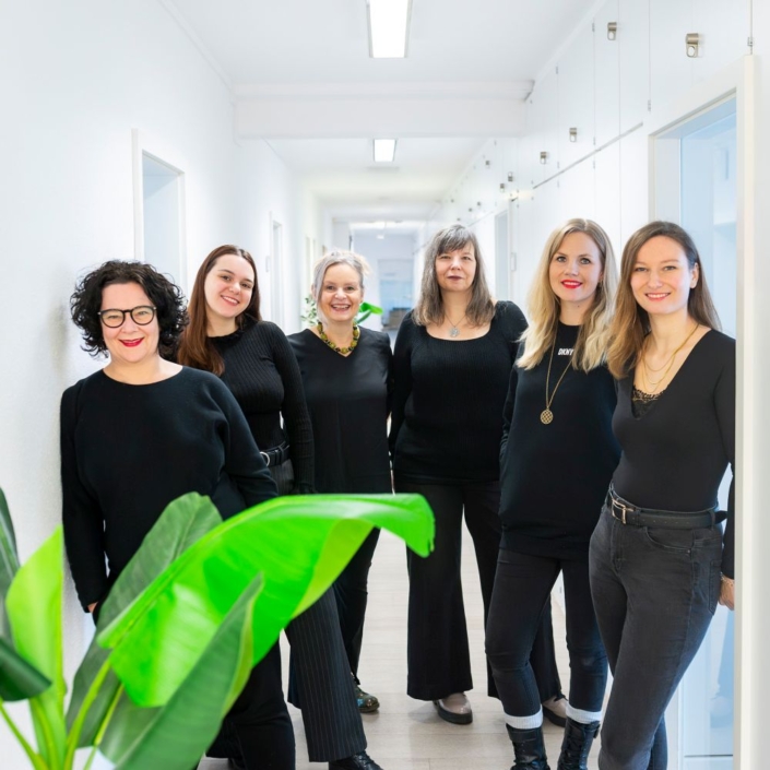 ©FotoAgenten_ALöffler_Lindmanns _214-I_1000 Sechs Frauen stehen lächelnd in einem hellen Flur mit weißen Wänden, gekleidet in schwarze Outfits. Auf der linken Seite ist eine große Grünpflanze zu sehen, die den professionellen Geist einer Event-Agentur, die sich auf die Betreuung von Online-Events spezialisiert hat, perfekt einfängt.