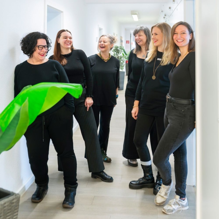 Team lacht Sechs meist schwarz gekleidete Frauen stehen in einem hellen Flur neben einer großen Grünpflanze und lächeln sich an, sie wirken entspannt und glücklich.